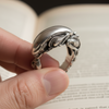 Silver alien ring with intricate design held by a hand against a blurred book background